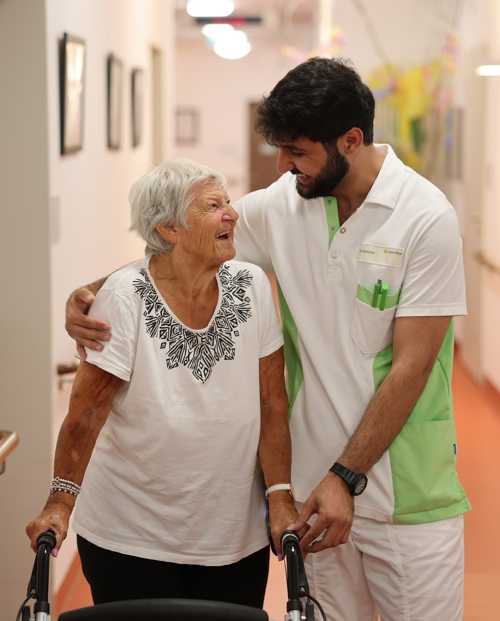 Pflegekraft begleitet eine ältere Bewohnerin mit Rollator durch den Flur. Beide lächeln sich herzlich an, was Wärme und Vertrauen vermittelt.