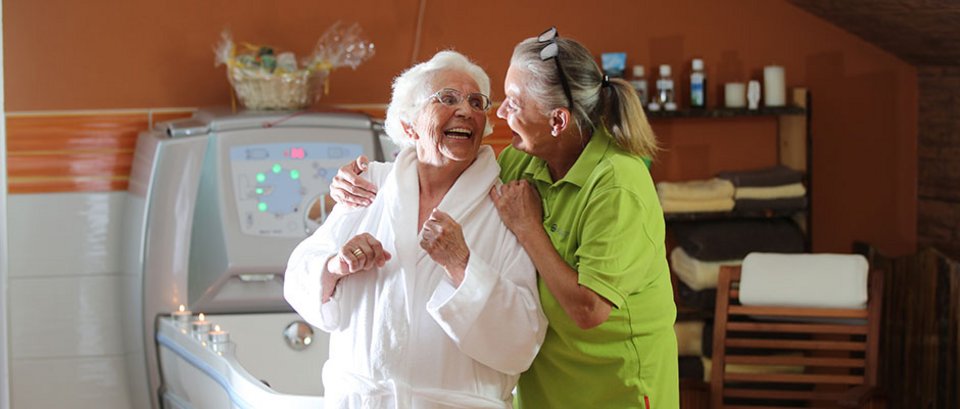 Eine Mitarbeiterin der AllgäuPflege und eine Bewohnerin sitzen lachend auf dem Badewannenrand