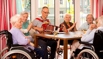 Seniorinnen sitzen an einem Tisch in der stationären Einrichtung der AllgäuPflege in Immenstadt, ein Mann spielt Klavier