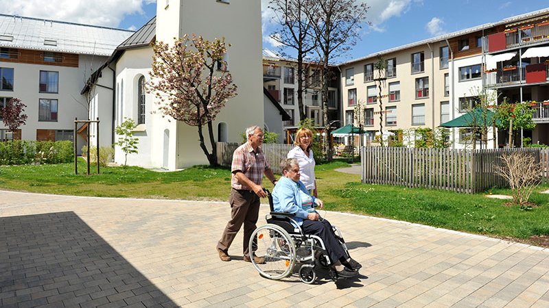 Ein Mann schiebt eine Frau im Rollstuhl durch den Garten des Spitals Sonthofen