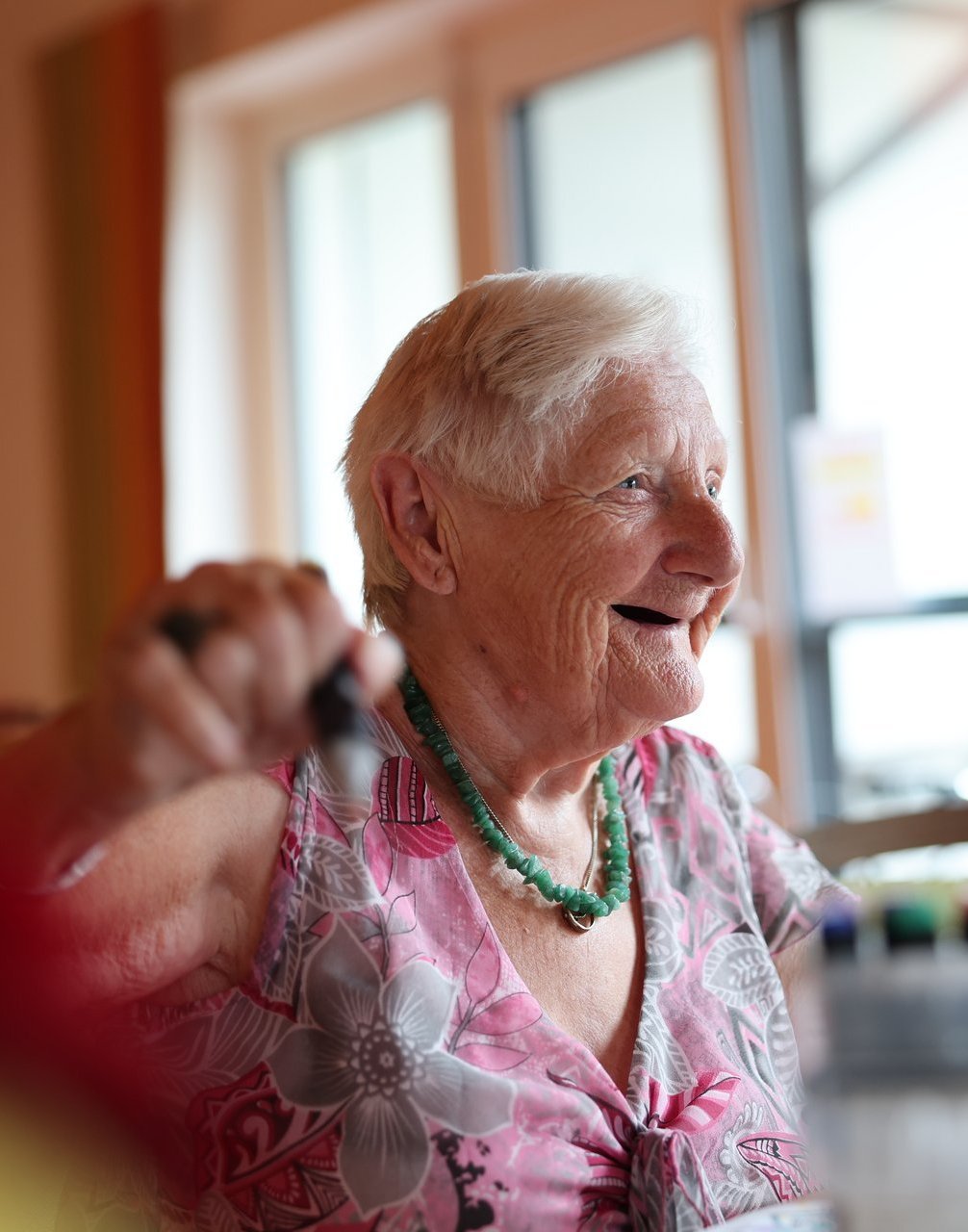 Ältere Frau sitzt lachend am Tisch im Gemeinschaftsraum des Spitals Immenstadt und malt. Sie trägt eine bunte Bluse und eine Halskette – Sinnbild für Lebensfreude im Alter.
