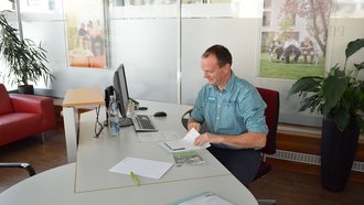 Ein Mann sitzt am Schreibtisch vor einem Bildschirm und sichtet Papierunterlagen.