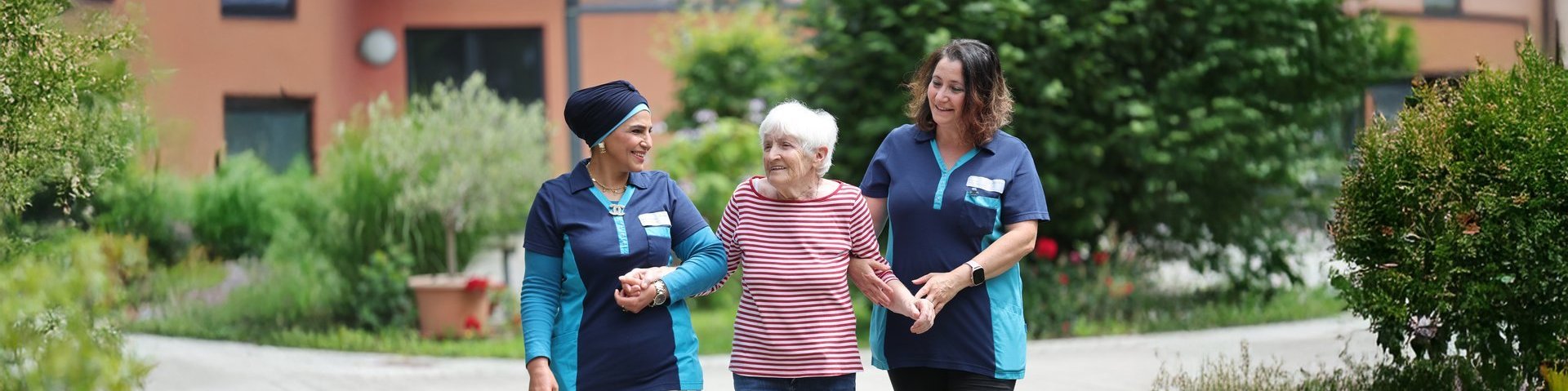 Zwei Pflegekräfte gehen mit einer älteren Frau Hand in Hand durch den Garten des Spitals Immenstadt. Die grüne Umgebung vermittelt Ruhe, Natur und Lebensqualität im Alter.