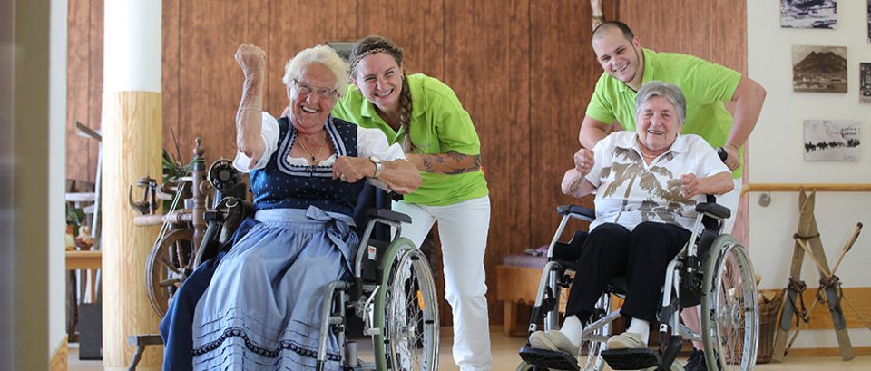 Zwei Frauen im Rollstuhl werden lachend von Mitarbeiterinnen der AllgäuPflege geschoben