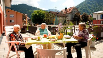Drei Senioren sitzen an einem Tisch auf einer Terrasse in der Sonne.