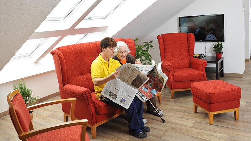 Eine Mitarbeiterin der AllgäuPflege liest einer Bewohnerin aus der Zeitung vor